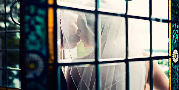 Beautiful photo by 2 Brides Photography of couple kissing behind window.